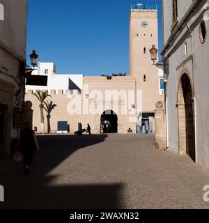 Strada nella storica medina con la torre dell'orologio davanti mentre qualcuno in silhouette spinge un carro nella città di Essaouira, in Marocco. 7 gennaio 2024 Foto Stock