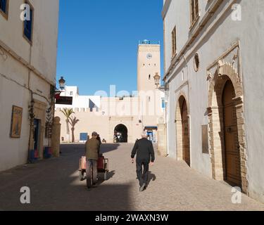 L'uomo spinge un carro in una strada nella storica medina con la torre dell'orologio davanti alla città di Essaouira, in Marocco. 7 gennaio 2024 Foto Stock