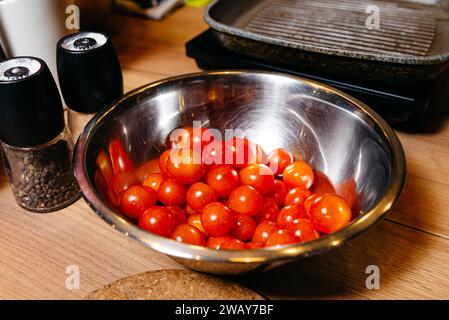 Pomodori ciliegini freschi in una ciotola riflettente in acciaio inossidabile su un piano di cucina in legno, con macinacaffè e shaker per sale nelle vicinanze. Foto Stock