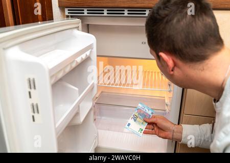 Baviera, Germania - 6 gennaio 2024: Un uomo con banconote in euro davanti a un frigorifero vuoto. Costi e prezzi per il concetto alimentare *** Ein Mann mit Euro Geldscheinen vor einem leeren Kühlschrank. Kosten und Preise für Lebensmittel Konzept Foto Stock