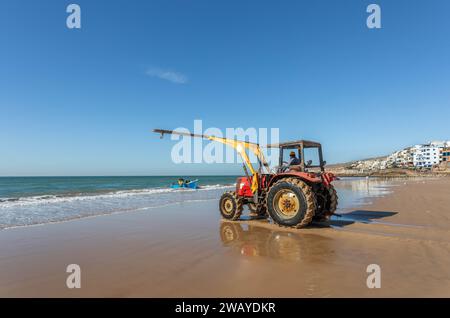 Un trattore è sulla spiaggia, pronto a sollevare un piccolo peschereccio di legno blu dall'acqua dopo una battuta di pesca. Taghazout, Marocco, Nord Africa. Foto Stock