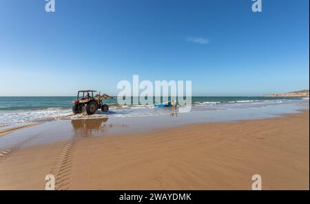 Un trattore è sulla spiaggia, pronto a sollevare un piccolo peschereccio di legno blu dall'acqua dopo una battuta di pesca. Taghazout, Marocco, Nord Africa. Foto Stock