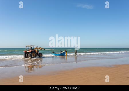 Un trattore è sulla spiaggia, pronto a sollevare un piccolo peschereccio di legno blu dall'acqua dopo una battuta di pesca. Taghazout, Marocco, Nord Africa. Foto Stock