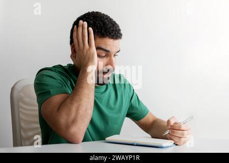 Uomo frustrato con la barba in una maglietta verde che tiene la testa con una mano Foto Stock