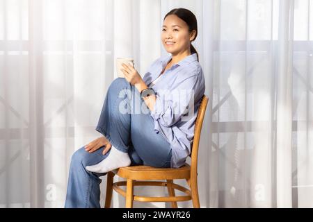 Felice attraente donna cinese seduta sulla sedia, bevendo tè Foto Stock