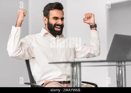 Entusiasta uomo d'affari indiano che festeggia il successo alla scrivania dell'ufficio Foto Stock