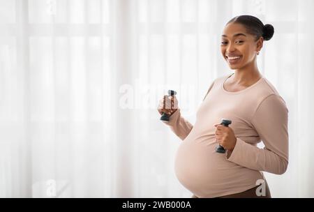 Donna nera incinta sorridente che si esercita con i campanelli a casa Foto Stock