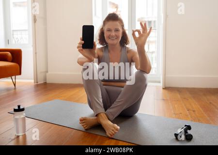 Donna anziana sportiva che tiene uno smartphone vuoto e mostra un gesto OK Foto Stock