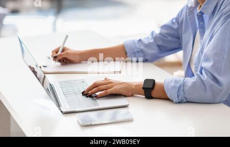 Donna professionista con una camicia blu multitasking, che digita su un notebook mentre scrive appunti Foto Stock