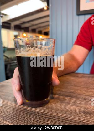 Un uomo che indossa una t-shirt rossa vivace tiene una pinta di birra scura su un tavolo di legno. Il portapacchi di cioccolato amaro freddo ha schiuma e bolle. Foto Stock