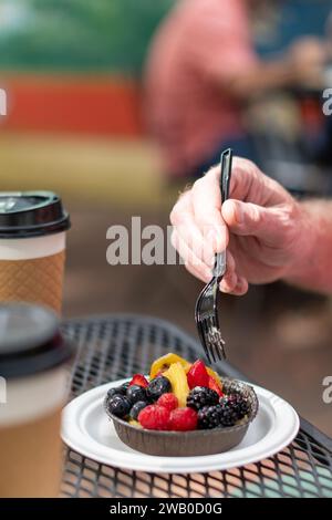 Una mano maschile che versa una forchetta in una pasta di frutta rotonda. La base è formaggio spalmabile con glassa. Le bacche sono lampone, fragola, mirtillo, ananas Foto Stock