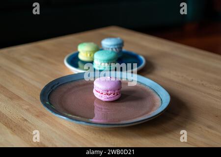 Quattro macaron francesi: Rosa, verde, blu e giallo. Gli eleganti dessert dolci assortiti in stile panino sono su un piatto blu e marrone e un tavolo di legno. Foto Stock