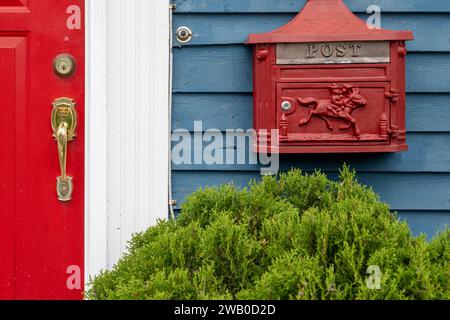 Sulla parete esterna di una casa in legno blu con una porta rossa vivace è fissata una cassetta postale in metallo rosso retrò, o letterbox. Foto Stock