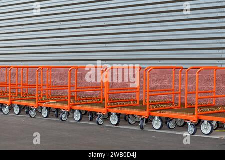 Una fila di carrelli per carichi pesanti in acciaio inossidabile di colore arancione è conservata all'esterno di un punto vendita con una parete esterna in metallo grigio. Una fila di buggy con caddy a spinta. Foto Stock