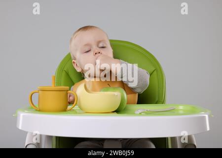 Piccolo bambino carino che mangia cibo sano in seggiolone su sfondo grigio Foto Stock