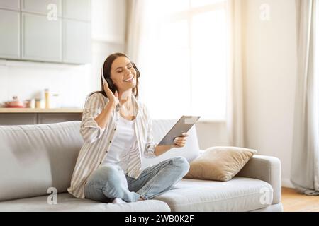 Donna gioiosa seduta a gambe incrociate su un divano, ascoltando musica tramite cuffie wireless Foto Stock
