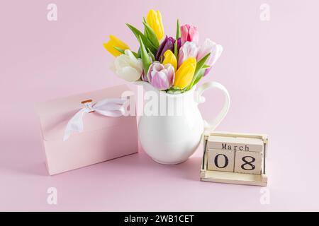 Bouquet di tulipani primaverili in brocca bianca su sfondo rosa accanto a una scatola per le vacanze e un calendario in legno con la data dell'ottavo marzo Foto Stock