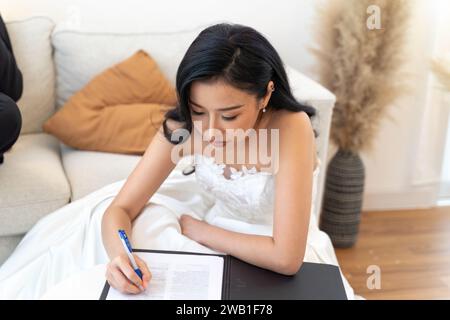 Donna asiatica in elegante abito da sposa che completa i documenti legali del matrimonio. Sposa con un abito bianco firma documenti di matrimonio, un momento ufficiale di impegno e bellezza Foto Stock