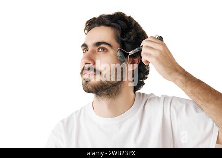 Il ritratto ravvicinato dell'uomo si muove agevolmente sulla pelle del viso con il rullo massaggiante su sfondo bianco dello studio. Concetto di routine per la cura del viso. Foto Stock