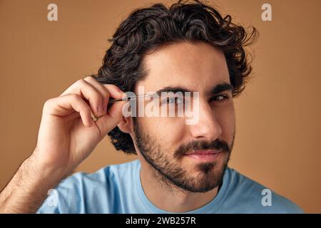 Ritratto ravvicinato di un giovane uomo attraente con pinzette che reggono i capelli brunette mentre modellano le sopracciglia sullo sfondo dello studio beige. Foto Stock