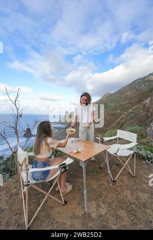 Una tranquilla colazione di coppia che condivide un pasto sulla scogliera, con vista sull'oceano sotto un cielo parzialmente nuvoloso. Campeggio e picnic Foto Stock