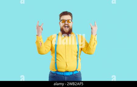 L'hipster dalla barba pazzo ed elegante si rallegra del suo successo mostrando il simbolo del rock and roll. Foto Stock