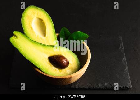 Avocado fresco maturo tagliato a metà in un recipiente di legno su un tabellone nero rustico. Concetto di alimentazione sana. Fotografia del cibo Foto Stock