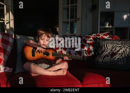 Il bambino in età prescolare siede sul divano e gioca con la chitarra per bambini alla luce del sole Foto Stock