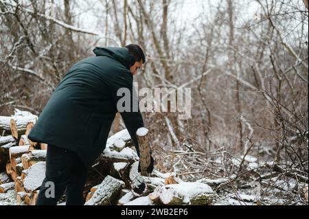 Uomo in abbigliamento invernale che raccoglie tronchi di legno e li accumula nella neve Foto Stock