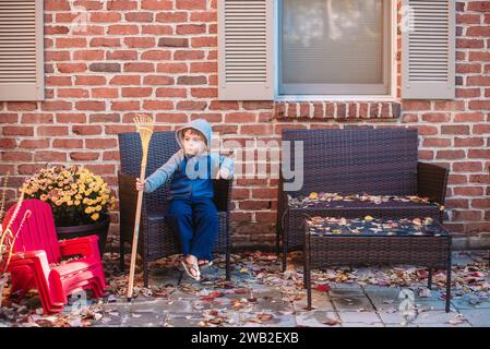 Il bambino in età prescolare con felpa con cappuccio si trova sul patio con un piccolo rastrello in autunno Foto Stock