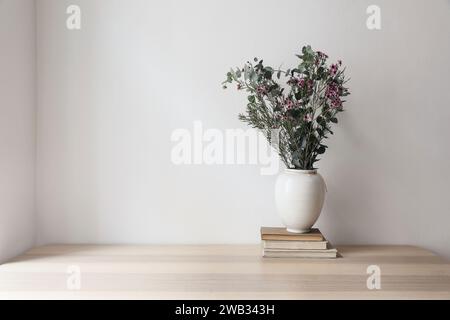 Vaso bianco con bouquet di eucalipto brnahces e fiori di cera. Vecchi libri su un tavolo di legno. Sfondo parete beige vuoto. Mockup neutrale Foto Stock