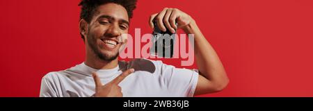 felice fotografo afro-americano che punta verso la fotocamera retrò e sorride su sfondo rosso, banner Foto Stock