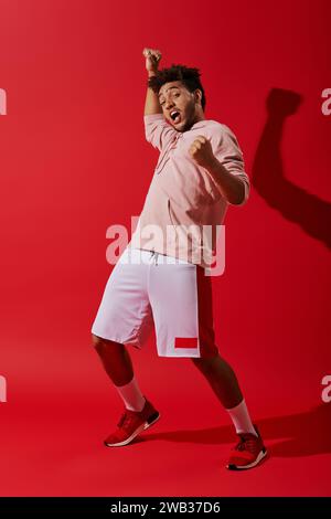 eccitato uomo afroamericano in abbigliamento sportivo rallegrato e guardando la fotocamera su sfondo rosso Foto Stock