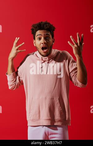 uomo afro-americano confuso in abiti da palestra gesturing con bocca aperta su sfondo rosso Foto Stock