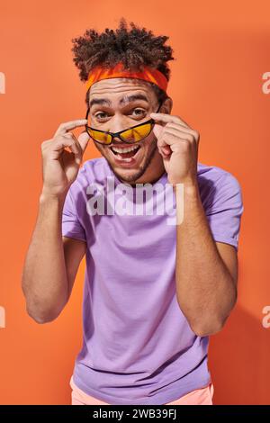 allegro uomo afro-americano con fascia sorridente e con occhiali da sole su sfondo arancione Foto Stock