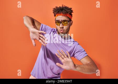 uomo afro-americano espressivo con occhiali da vista e cerchietto con gesti su sfondo arancione Foto Stock