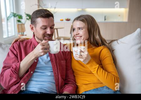 La coppia felice, il fidanzato e la fidanzata, bevono un caffè seduti sul divano di casa, godendosi la mattina a parlare Foto Stock