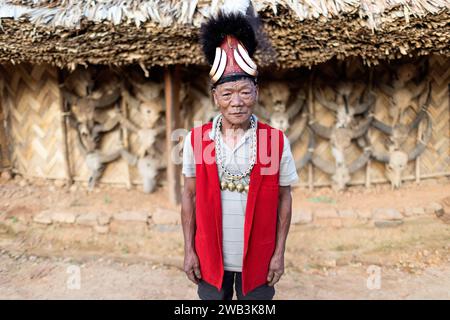 Anziano della tribù Konyak in abiti cerimoniali tradizionali, in piedi davanti a una casa in legno konyak, decorata con teschi di toro, Nagaland, India Foto Stock