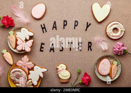 Sfondo pastello primaverile con testo Happy Easter. Biscotti pasquali decorati, fiori rosa, piume e uova distese Foto Stock