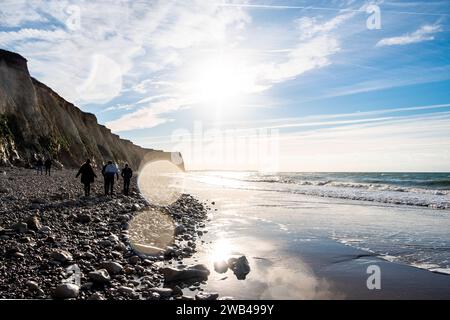 La fotografia raffigura un gruppo di persone che cammina lungo un tratto roccioso di una spiaggia con torreggianti scogliere su un lato. Il sole mattutino brilla, creando un effetto flare e proiettando le ombre degli escursionisti sulla spiaggia di ciottoli. Le onde incontrano dolcemente la spiaggia, contribuendo alla serena atmosfera mattutina. Le scogliere catturano la luce, mettendo in evidenza le loro texture e strati, che parlano della storia geologica della costa. L'immagine cattura la tranquillità di una passeggiata mattutina in riva al mare e l'interazione senza tempo tra terra, mare e cielo. Passeggiata lungo la costa sotto la domenica del mattino. Alto Foto Stock