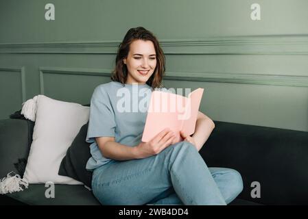 Gioiosa giovane millenaria che legge un libro mentre si rilassa su un divano in un accogliente soggiorno Foto Stock