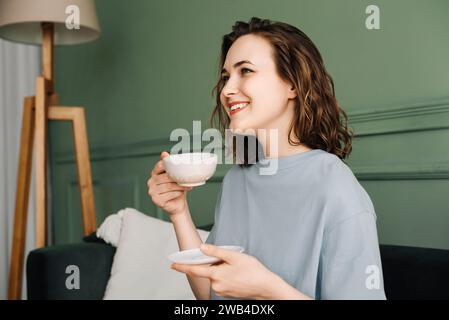 Rilassata donna di mezza età che gusta il tè sul divano e osserva il Copy Space. Sereno momento di Contentment. Donna a riposo, tazza di tenuta, spazio per testo o. Foto Stock