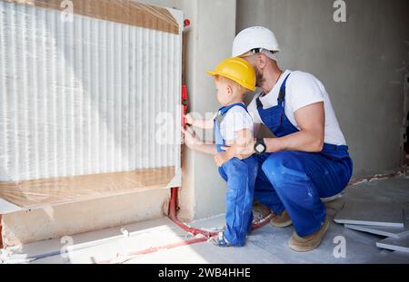 Padre e figlio piccolo che utilizzano una livella a bolla d'aria durante l'installazione del radiatore di riscaldamento nell'appartamento. Tubi di misurazione maschio per idraulici e bambini a livello con strumento edile. Foto Stock