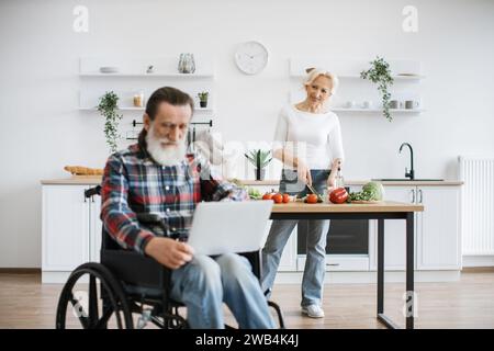 Bella moglie caucasica prepara insalata sana in cucina mentre suo marito vecchio con la barba grigia siede in sedia a rotelle e lavora sul computer portatile. Foto Stock