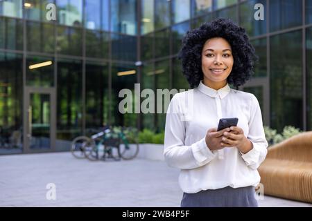 Ritratto di una giovane donna d'affari afroamericana che si trova fuori da un ufficio, che tiene un telefono cellulare e sorride davanti alla fotocamera. Foto Stock