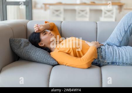 Donna indiana malata sdraiata sul divano, tenendo stomaco e testa Foto Stock