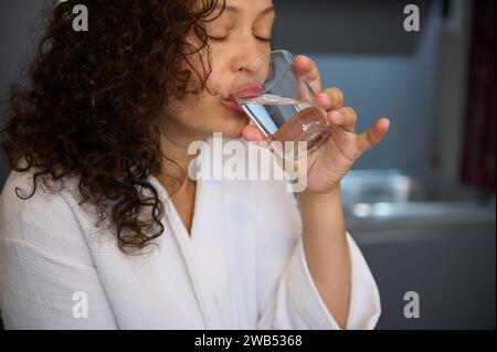 Ritratto in primo piano di una bella donna multietnica dai capelli ricci, vestita con accappatoio bianco, sorseggiando acqua minerale mentre prendeva medicine, un dolore Foto Stock