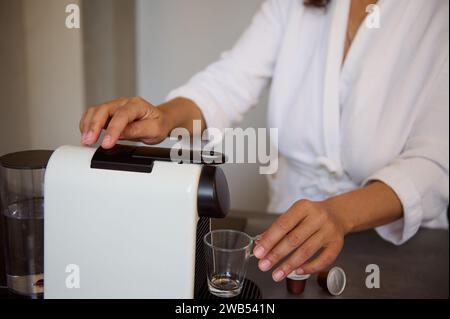 Attenzione selettiva a una macchina da caffè a capsule per uso domestico e donna che prepara caffè fresco al mattino per la colazione, accanto al bancone della cucina. Italiano Foto Stock