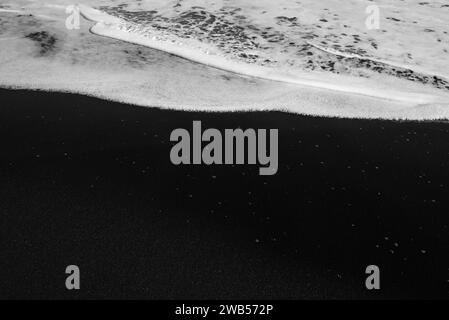 Spiaggia di sabbia nera per lo più sfocata con schiuma bianca di onde marine sullo sfondo Foto Stock