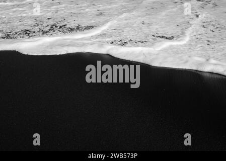 Spiaggia di sabbia nera per lo più sfocata con schiuma bianca di onde marine sullo sfondo Foto Stock